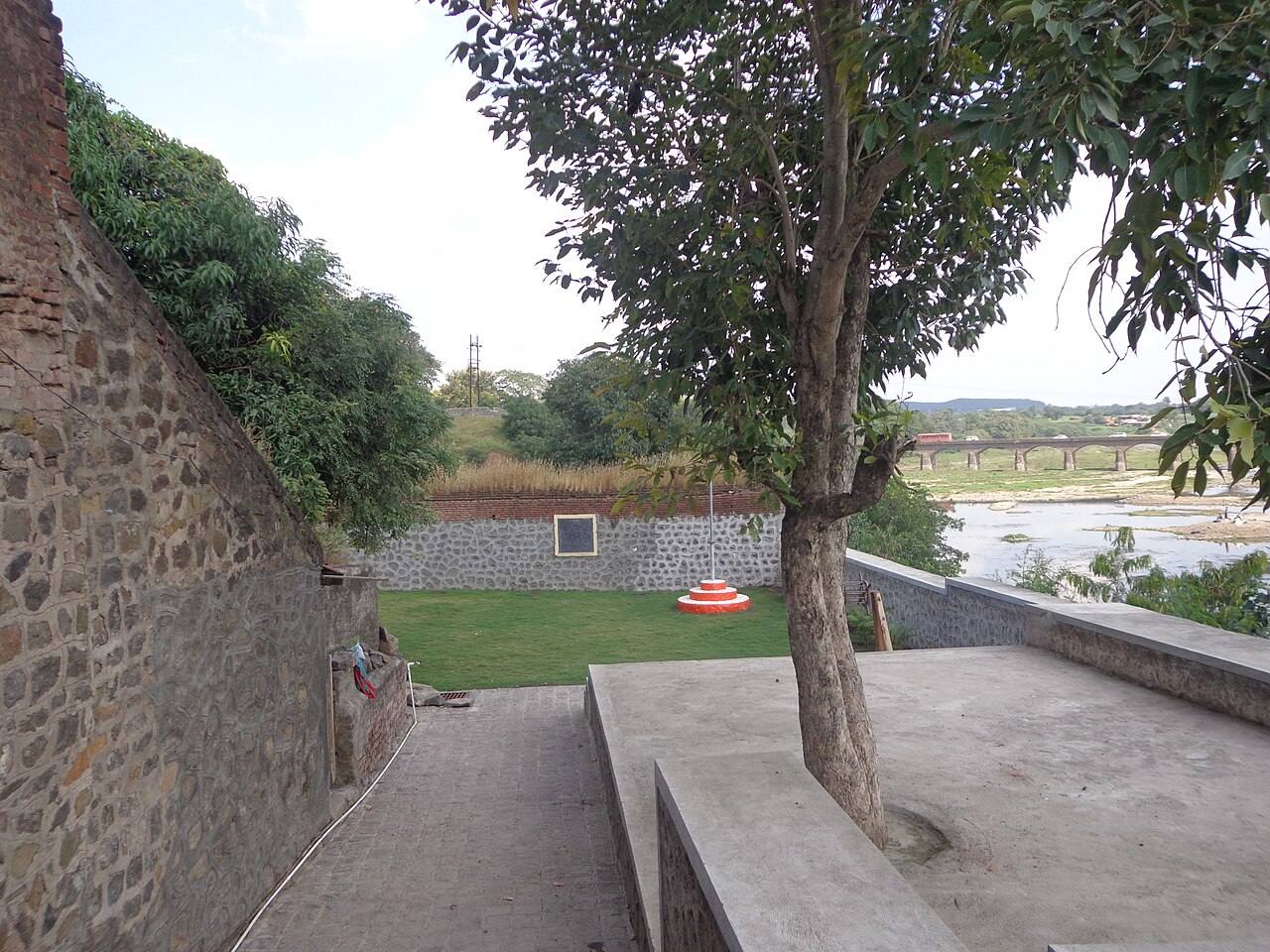 The Rajguru Wada site at Rajgurunagar today: stone perimeter walls enclosing a lawn with a flagstaff base and a commemorative plaque, a tree in the foreground, and the Bhima river with a road bridge beyond